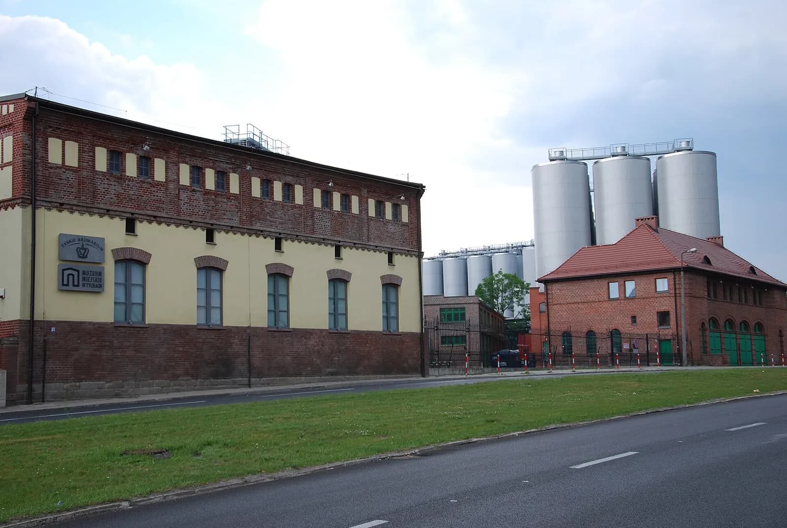 A long brick and yellow building with many small windows stands next to a road, with tall silver industrial silos and another brick building with a red roof in the background under a cloudy sky.